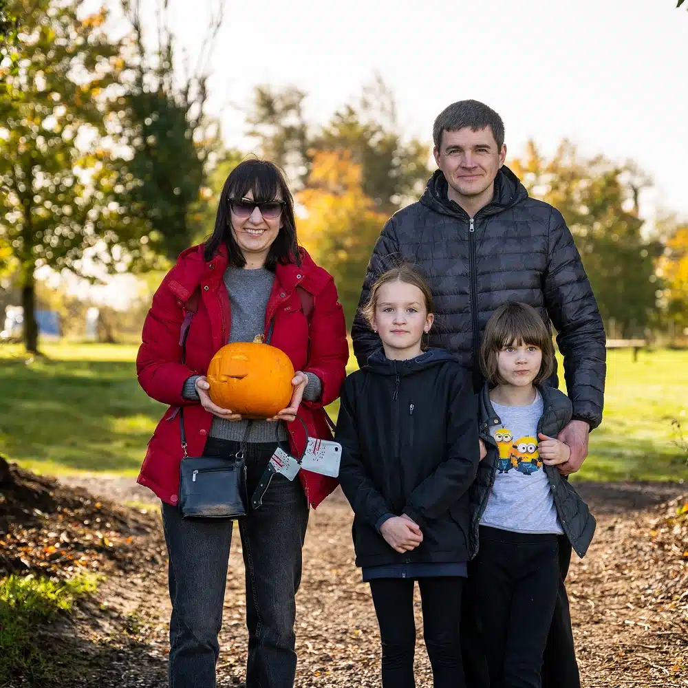 A family of four stands outdoors on a sunny day. The woman holds a carved pumpkin, and everyone is wearing jackets. The background features green grass and trees with autumn foliage.
