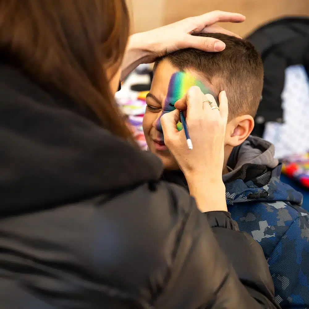 A child with closed eyes is having their face painted with a colourful rainbow design by an artist, who is gently holding the child's head for support.