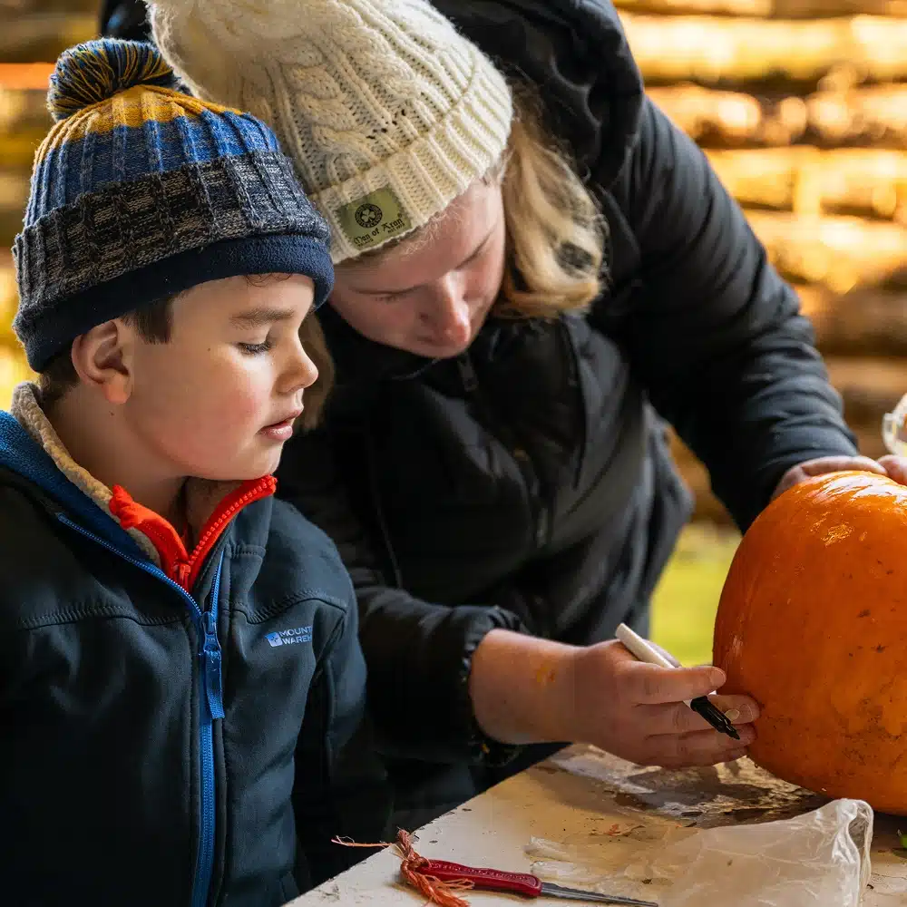 An adult in a white knitted hat helps a child in a blue hat draw on a pumpkin with a black marker at a table, preparing for pumpkin carving. They are both bundled in jackets, suggesting a chilly day.