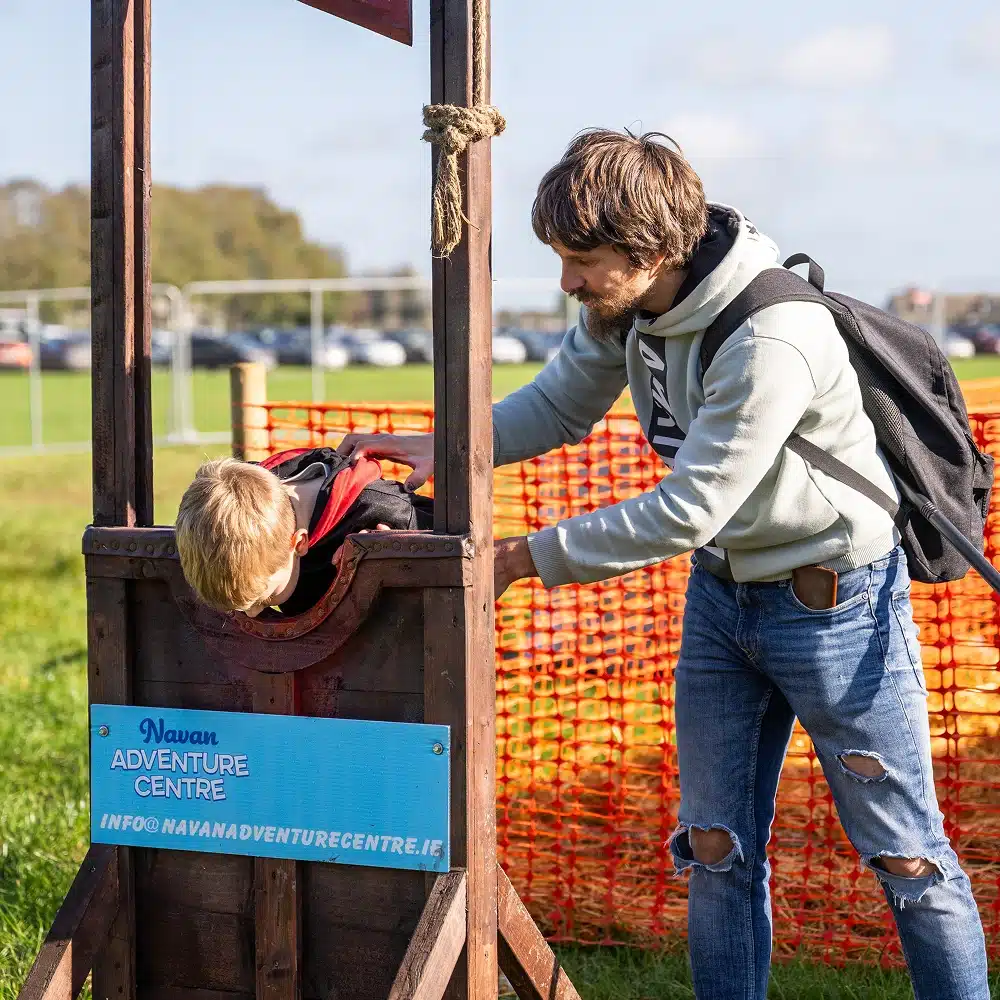 A young boy with his head and hands in wooden stocks at Navan Adventure Centre, while an adult with a rucksack helps him. Orange safety netting and parked cars are visible in the background on a sunny day.