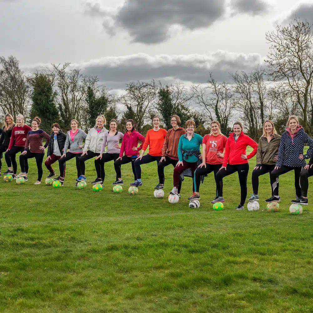 A group of thirteen women, possibly enjoying Navan hen and stag parties, stand in a line on grass with one foot on a football, smiling in colourful casual clothes. Trees and a cloudy sky form the backdrop.