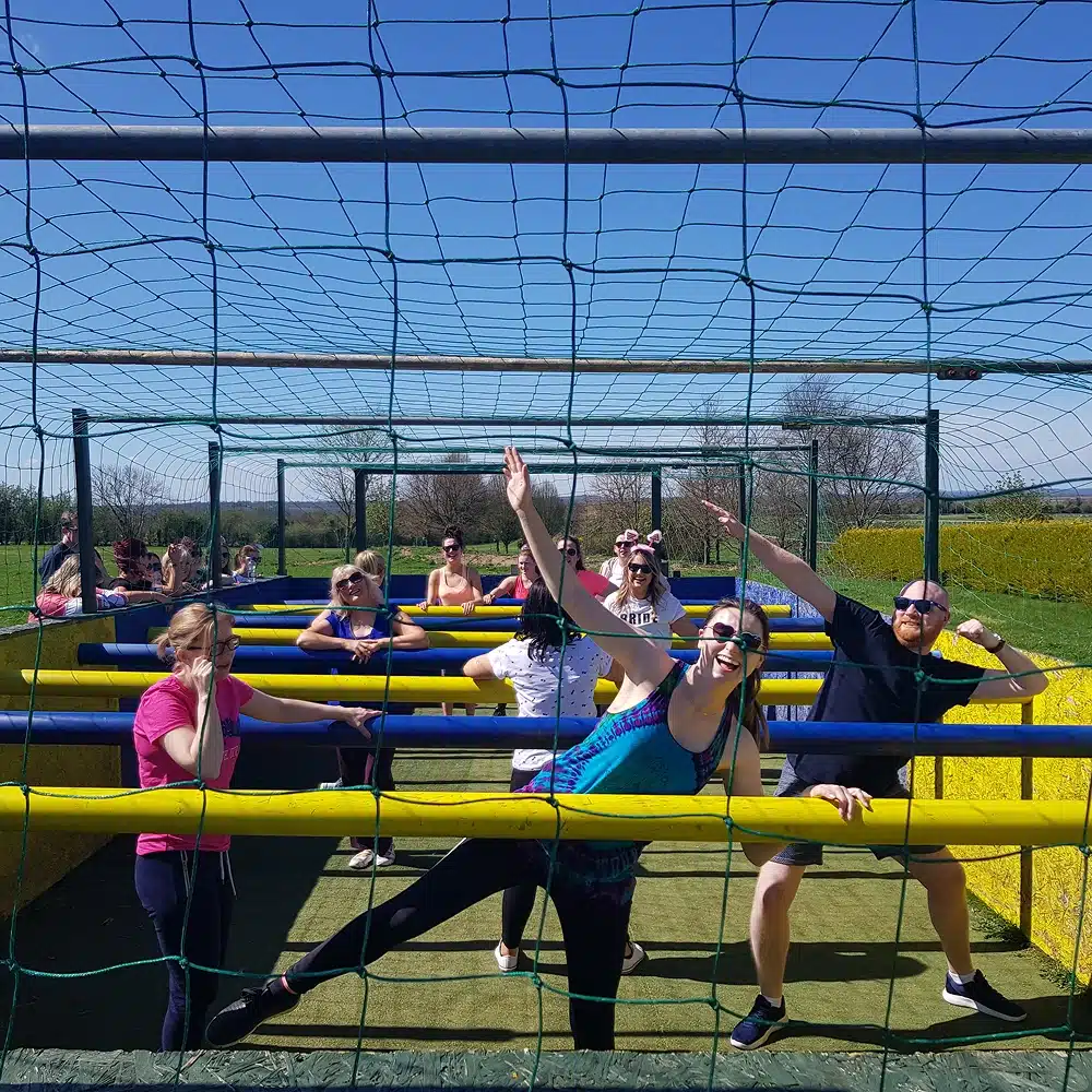 A group of people outdoors, possibly celebrating Navan hen and stag parties, play human table football on a grassy field under a net. They are posing joyfully, smiling and stretching their arms on a sunny day.
