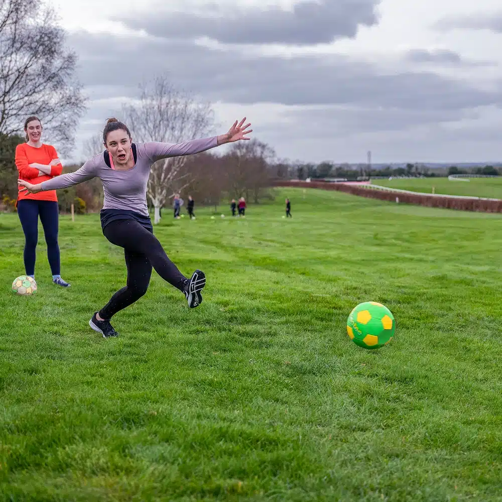 A woman kicks a brightly coloured football on a grassy field, with another woman watching nearby—an energetic scene perfect for Navan hen and stag parties. Several people are visible in the background under a cloudy sky.