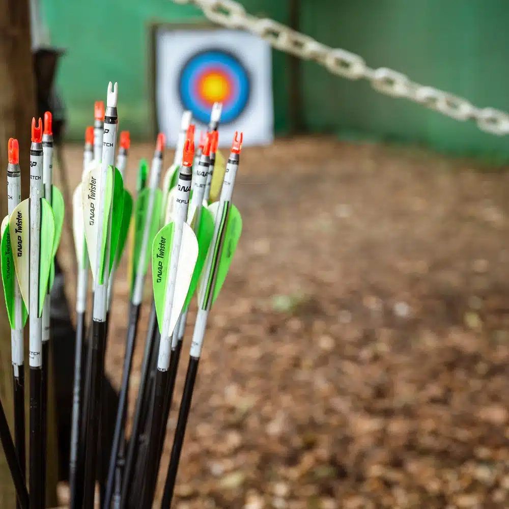 A bundle of arrows with green fletching sits in the foreground, whilst a colourful archery target—perfect for Navan hen and stag parties—appears blurred in the background outdoors. A white chain runs horizontally across the image.