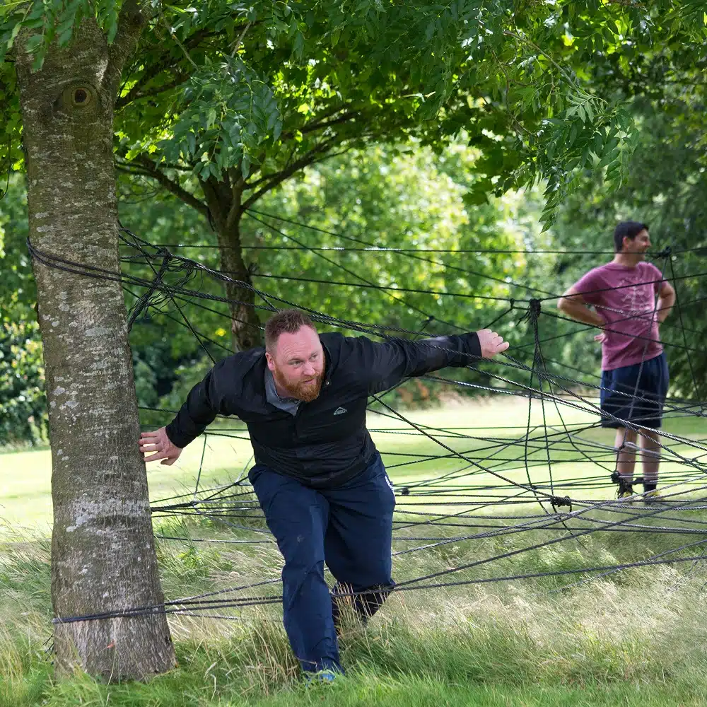 A man in dark clothing crouches and carefully navigates through a web of ropes strung between trees in a grassy park, while another man in a maroon shirt stands in the background—an activity popular at Navan hen and stag dos.