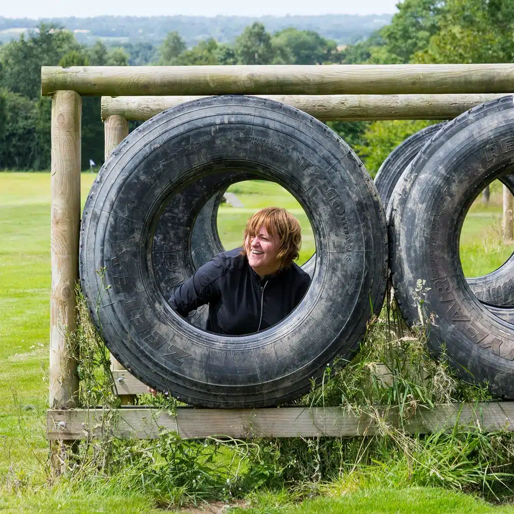 A woman with short hair and a black jacket crawls through a large tyre obstacle on a grassy outdoor course, smiling—an exciting challenge perfect for Navan hen and stag parties, with trees and fields in the background.
