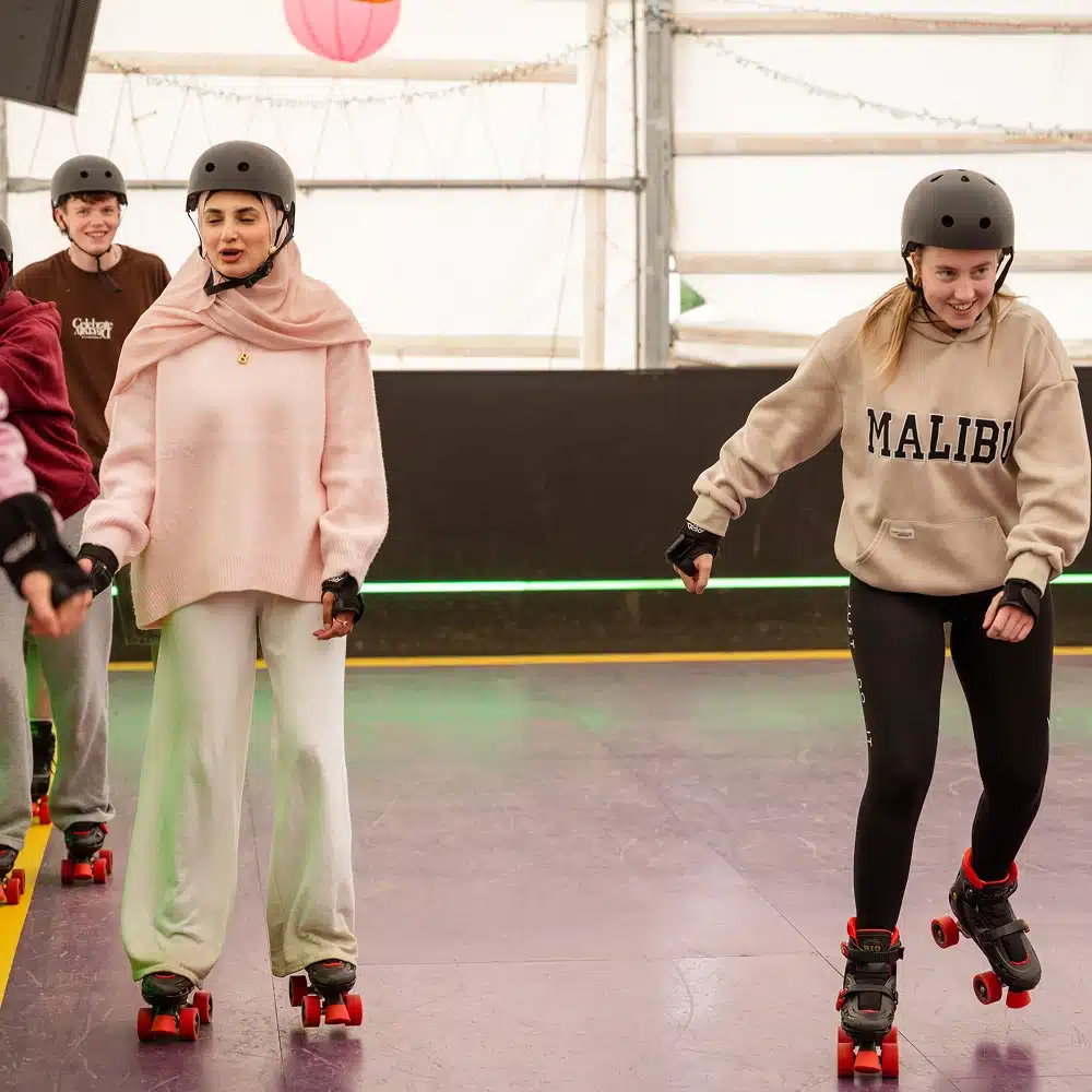 Four people roller skating indoors on a smooth, purple floor—perfect fun for Navan hen and stag parties. Wearing helmets and casual clothes, two hold hands, smiling as they enjoy themselves. The marquee-like wall and colourful decorations complete the scene.