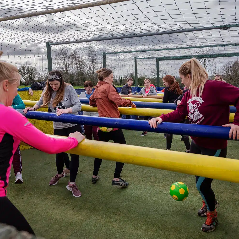 A group of women, celebrating Navan hen and stag parties, play human table football outdoors, each holding onto horizontal bars while kicking a green and yellow football on a fenced court under a cloudy sky.