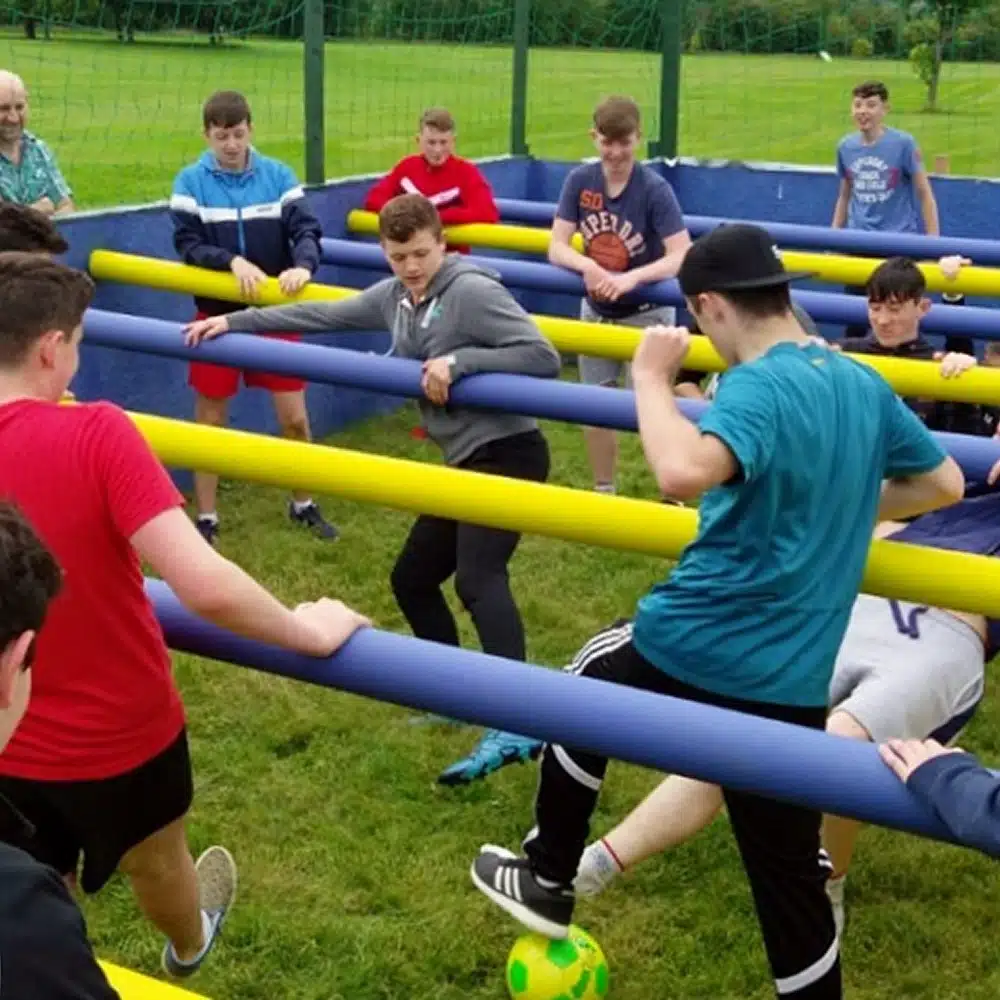 A group of teenagers play Navan human table football outdoors, each holding onto horizontal yellow and blue bars as they try to kick a green and yellow football on the grass.