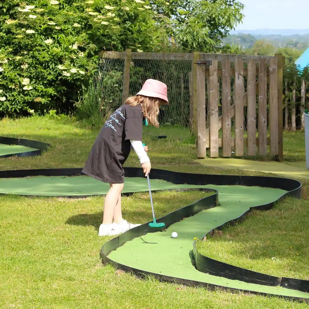 A young girl wearing a pink hat, black shirt, and white trainers plays crazy golf outdoors on a sunny day at a Navan bouncy castle party, preparing to hit a golf ball on a winding green course surrounded by grass and trees.