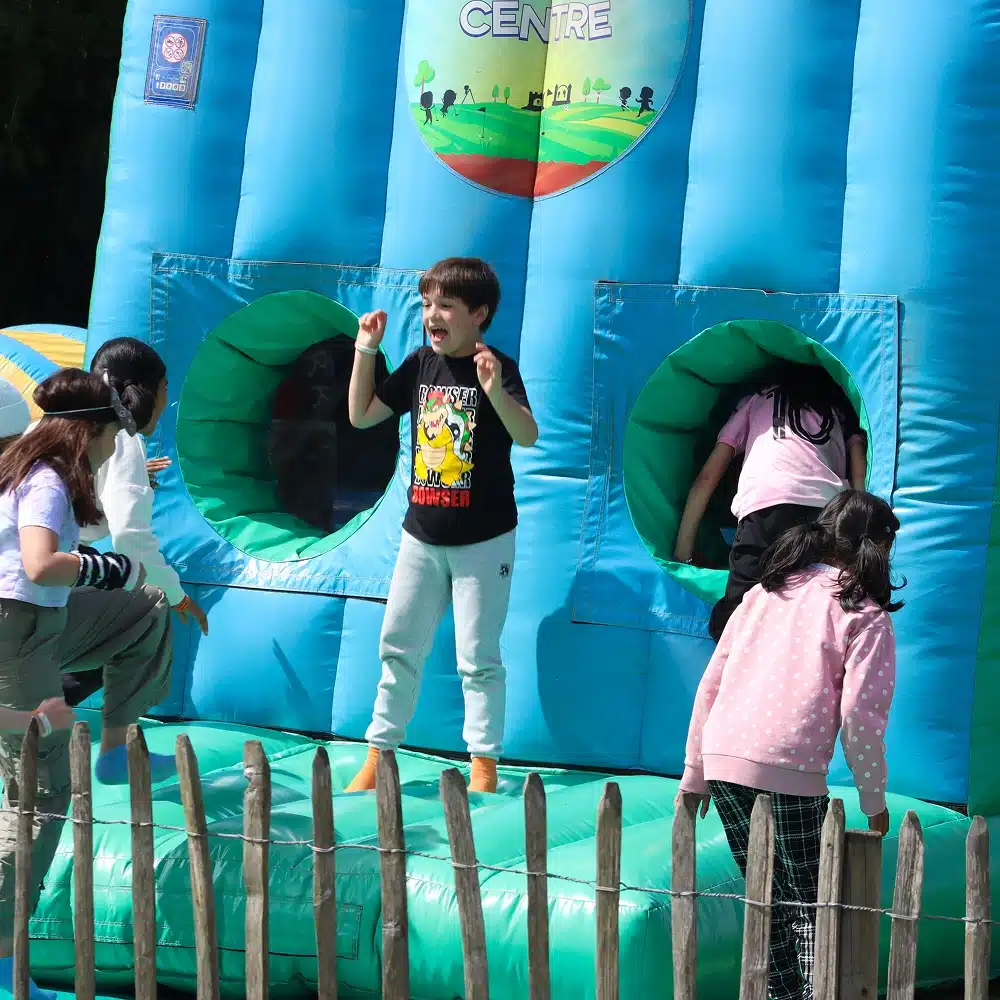 Several children play on a blue inflatable bouncy castle outdoors at NeuroFest Navan. A boy in a black T-shirt jumps in the centre while other kids climb and enter through circular openings. A wooden fence is visible in the foreground.