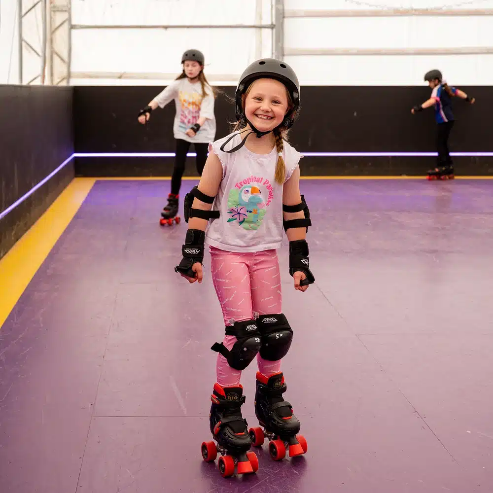 A young girl wearing a helmet and protective pads smiles whilst roller skating indoors at the Navan indoor roller disco. Two other children skate in the background on a purple and yellow skating rink.