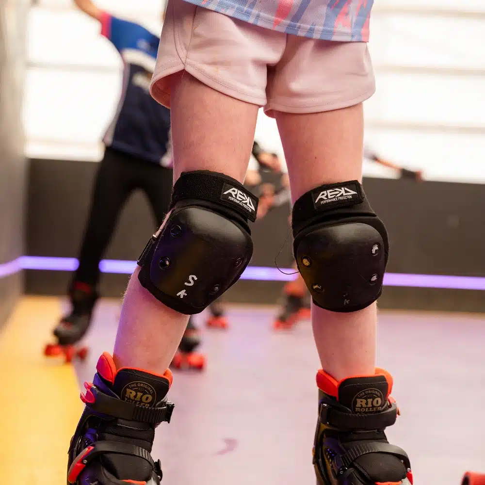 A close-up of a person wearing knee pads, shorts, and roller skates at the Navan indoor roller disco, standing on the rink with other skaters and a blurred background visible.