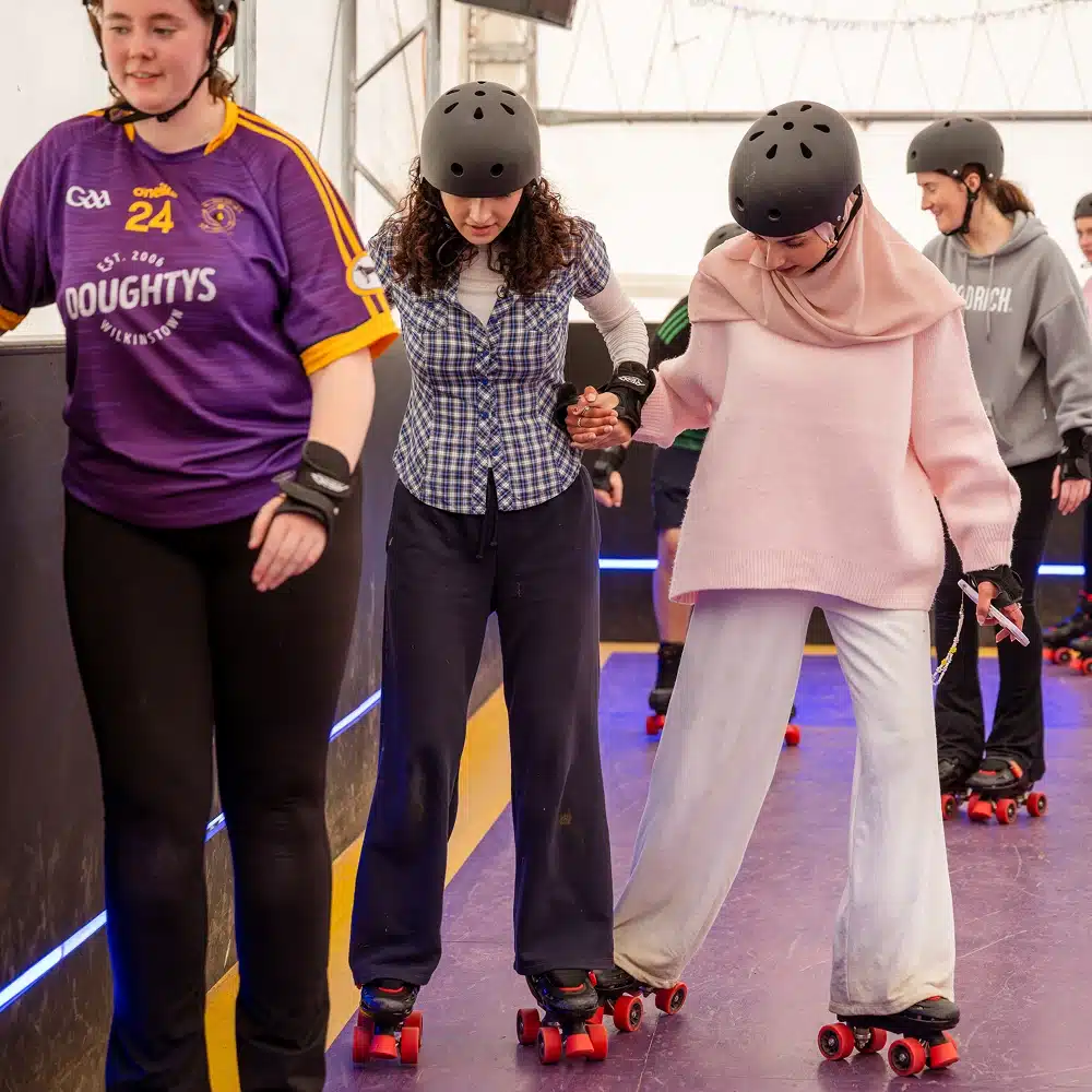 Four people wearing helmets and roller skates practise at the Navan indoor roller disco. Two women in the centre hold hands for balance, one in a chequered shirt and the other in a pink hijab and jumper, while others skate alongside them.