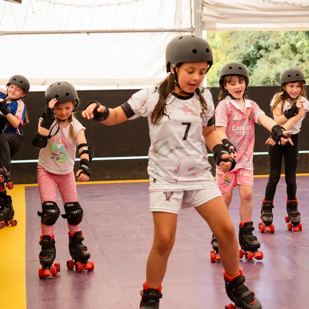 Five children wearing helmets and protective gear roller skate indoors at the Navan indoor roller disco. One child in white leads whilst others in pink follow, smiling and enjoying the activity. The background shows a partially open marquee and greenery outside.