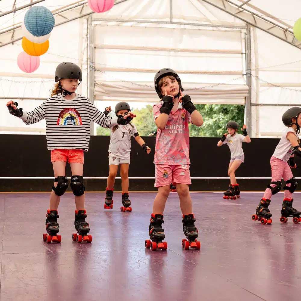Five children wearing helmets and pads roller-skate indoors on a purple floor at the Navan indoor roller disco, gliding under colourful hanging lanterns. Two girls in front pose with their arms out as three others skate in the background.