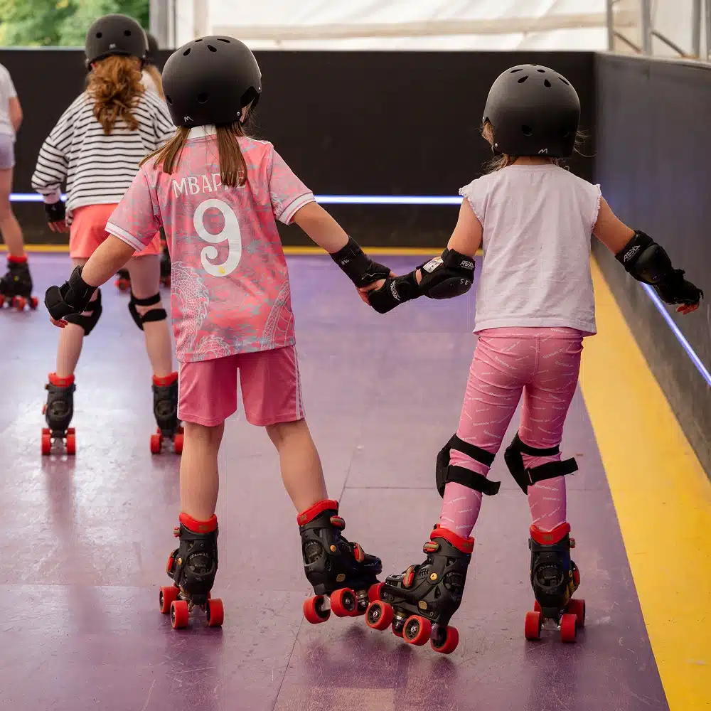 Several young children wearing helmets, knee pads, and roller skates hold hands whilst skating at the Navan indoor roller disco. The child in the centre wears a pink jersey with "MBAPPE 9" on the back, gliding across the colourful rink.
