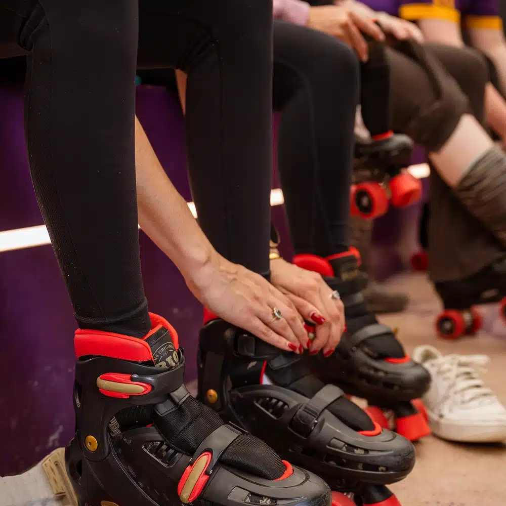 A close-up of people at the Navan indoor roller disco, sitting on a bench and putting on black and red roller skates. Their hands are adjusting the skates, with some wearing leggings and one person’s trainers beside them.