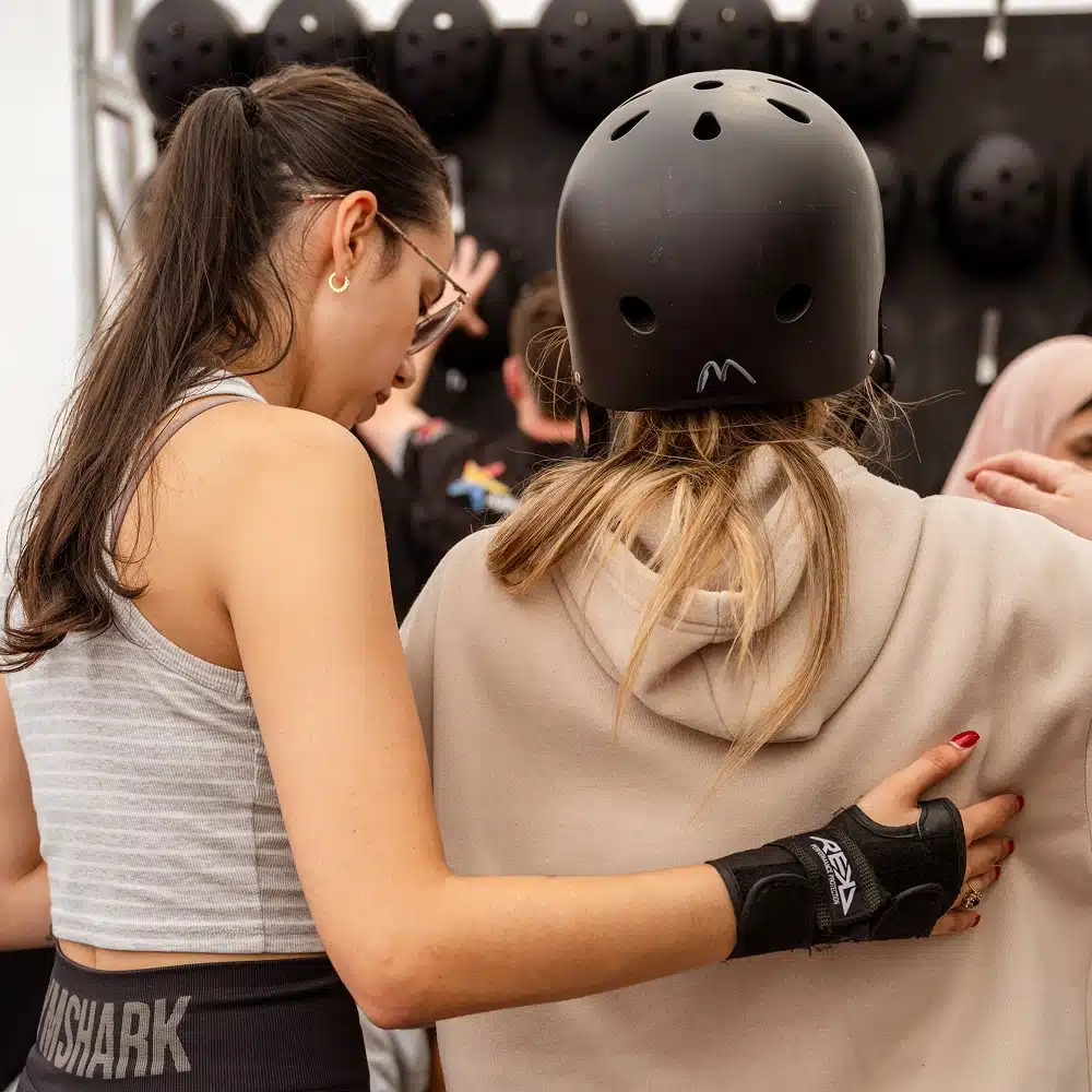 Two women stand close together at the Navan indoor roller disco, one with her arm supportively round the other. One wears a sports bra and glasses; the other has a helmet and beige hoodie. Helmets hang on a rack in the background.