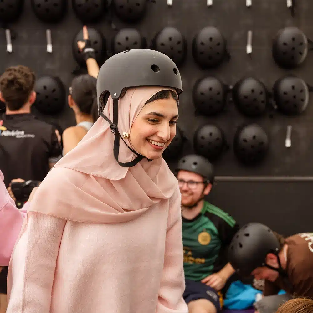 A smiling woman in a pink hijab and jumper wears a black helmet at the Navan indoor roller disco. Other people with helmets are in the background, with numerous black helmets hanging on a dark wall.
