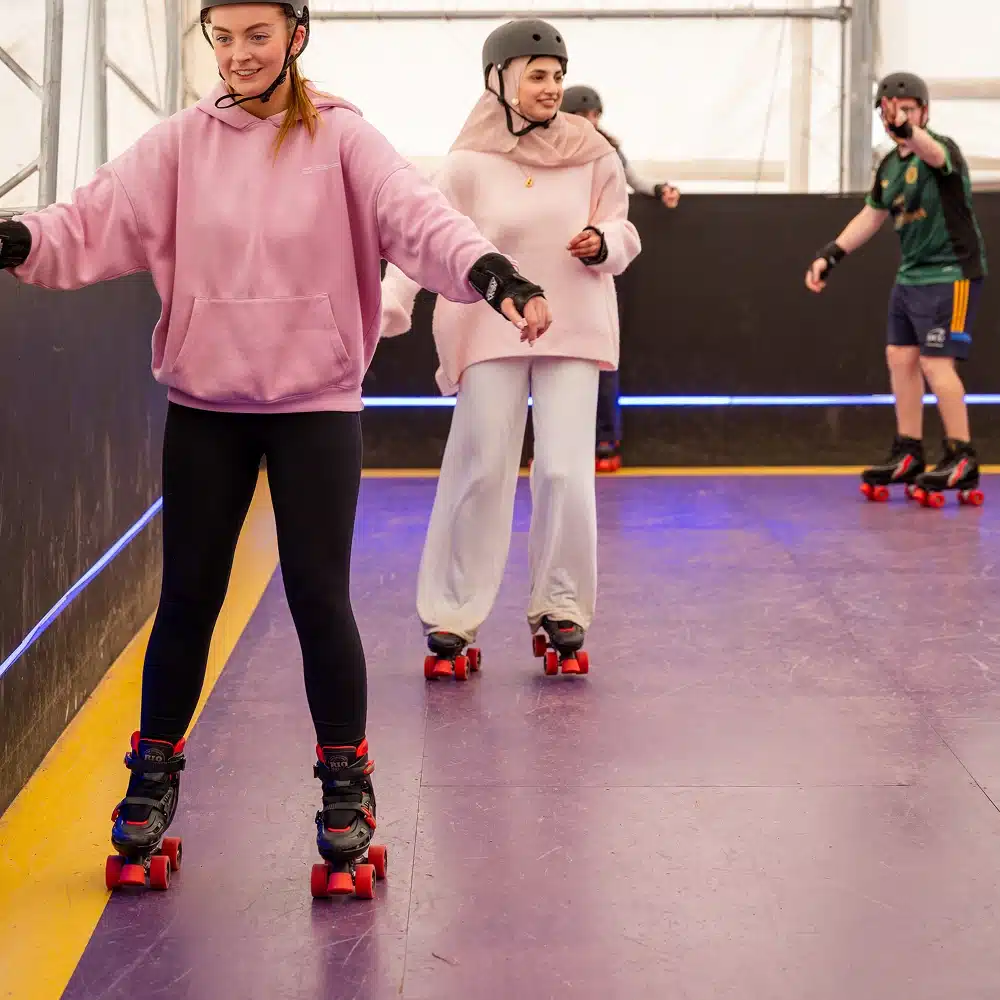 Two young women in helmets and casual clothes roller skate indoors at the Navan indoor roller disco, smiling and balancing, whilst two other skaters enjoy the vibrant purple and yellow rink in the background.