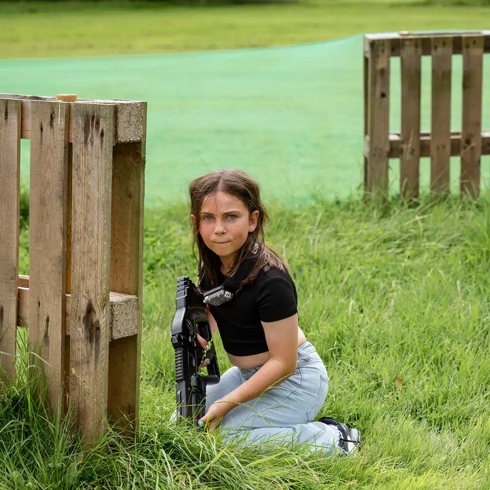 A young girl with long brown hair, wearing a black t-shirt and light blue trousers, kneels on grass whilst holding a laser tag gun beside a wooden barrier at Navan outdoor laser tag.