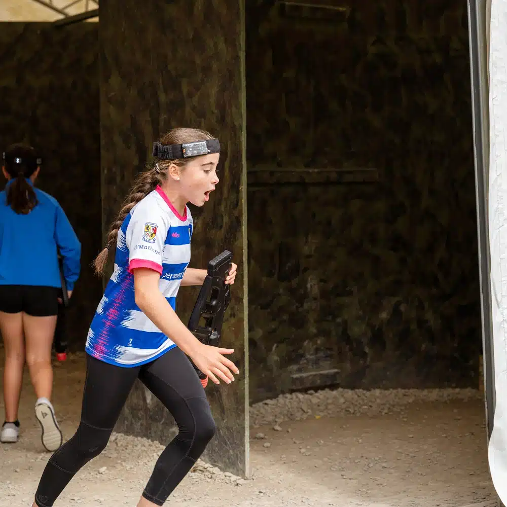 A young girl wearing a headband and sporty clothes runs excitedly with a laser tag gun at Navan outdoor laser tag. Two other children are visible in the background, walking away inside a dimly lit, camouflaged play area.