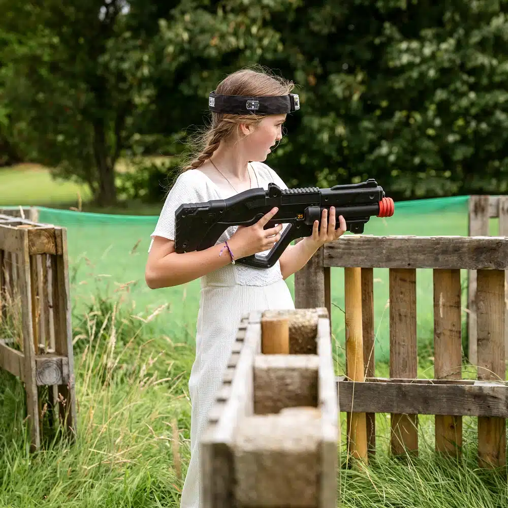 A girl in a white dress holds a large black toy laser gun and wears a sensor headband, standing outdoors near wooden barriers and tall grass, ready for an exciting game of Navan outdoor laser tag.