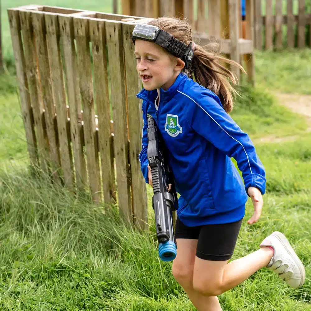 A young girl wearing a blue jacket and headband runs outside on grass, smiling and holding a toy laser tag gun at a Navan outdoor laser tag event, with a wooden structure in the background.