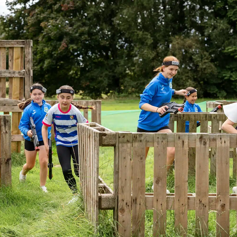 Children and a young adult, all wearing headbands and holding laser tag guns, run through a wooden outdoor obstacle course on a grassy field at Navan outdoor laser tag. Trees are visible in the background.