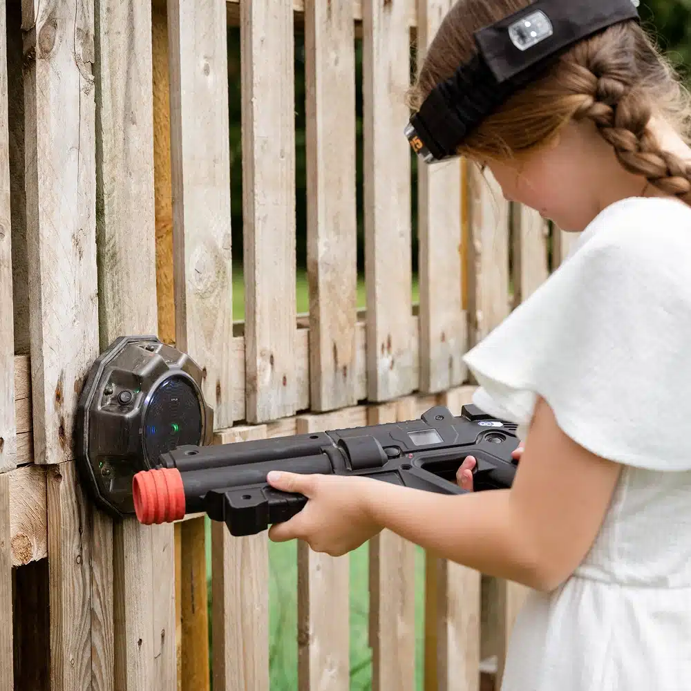 A girl with plaited hair, wearing a white dress and a black headband with sensors, enjoys Navan outdoor laser tag, aiming her toy laser gun at a sensor mounted on a wooden fence.