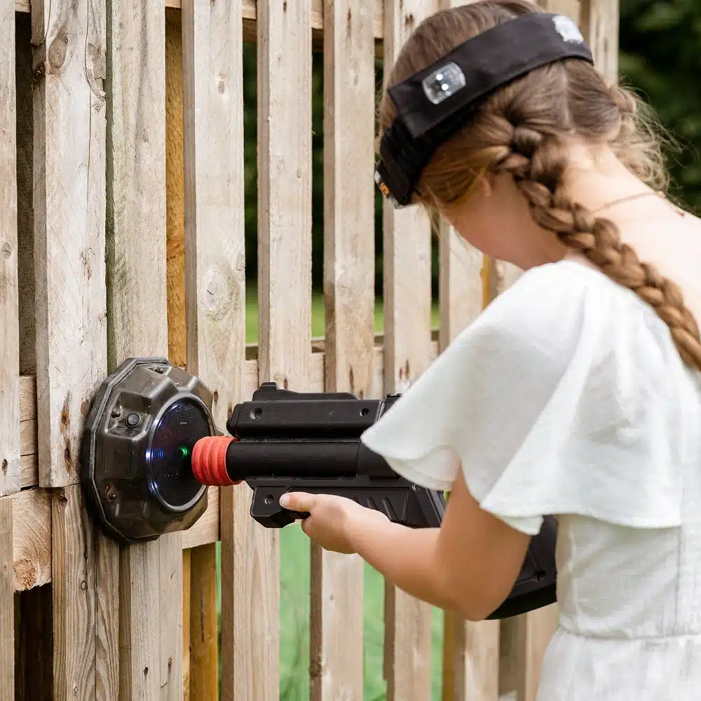 A girl with a plait and a hairband aims a black toy blaster at a target attached to a wooden fence outdoors during Navan outdoor laser tag. She is wearing a white dress.