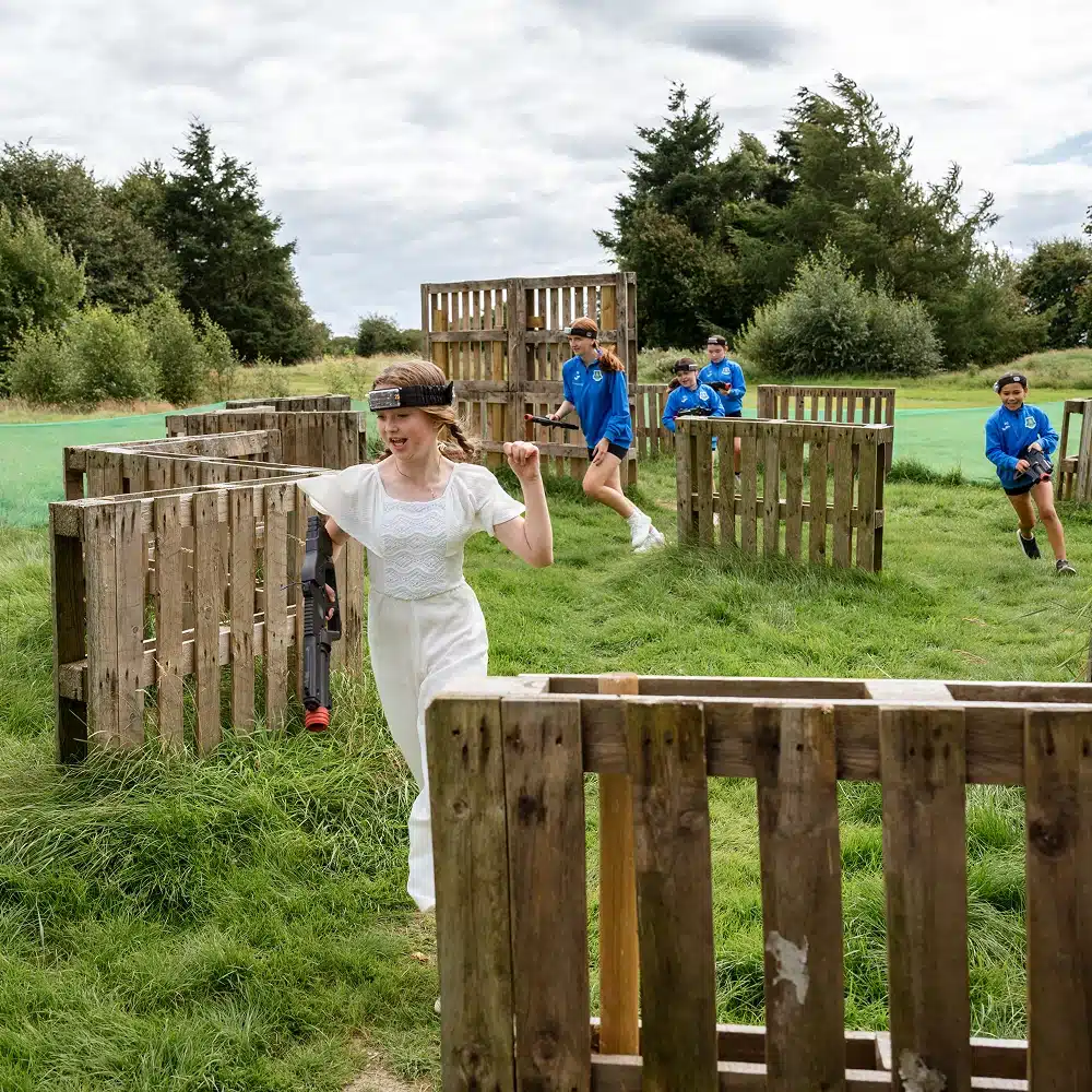 A group of children play Navan outdoor laser tag in a grassy area with wooden pallet barriers. One child in a white outfit runs ahead, while others in blue shirts follow, aiming their laser guns. Trees and cloudy sky are in the background.
