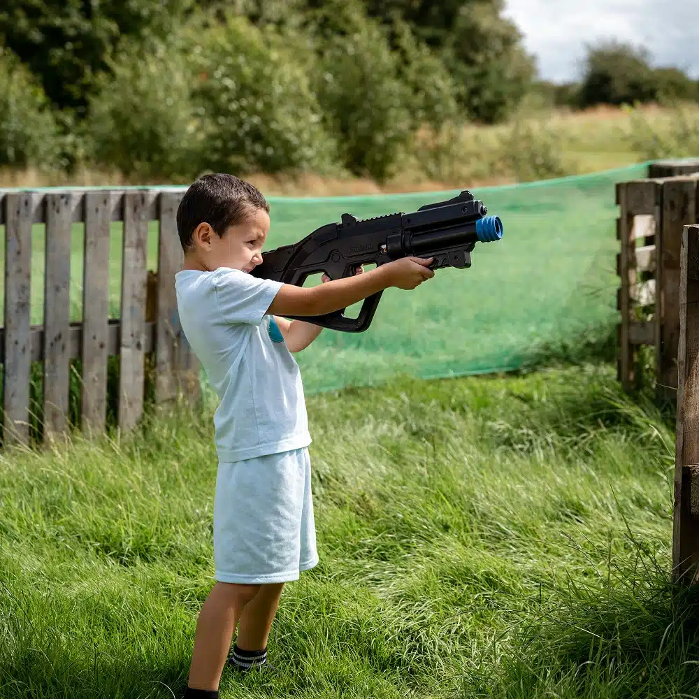 A young boy in light clothing stands on grass outdoors, holding and aiming a large toy gun with a blue tip—ready for action at a Navan laser tag party. Wooden fences and green netting are visible in the background.