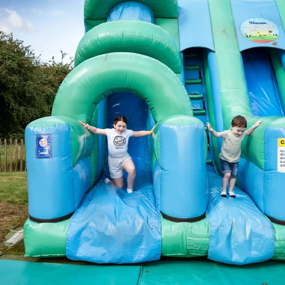 Two children, a girl and a boy, are playing and smiling as they come down a blue and green inflatable slide outdoors on a sunny day during a Navan laser tag party. Trees and grass are visible in the background.