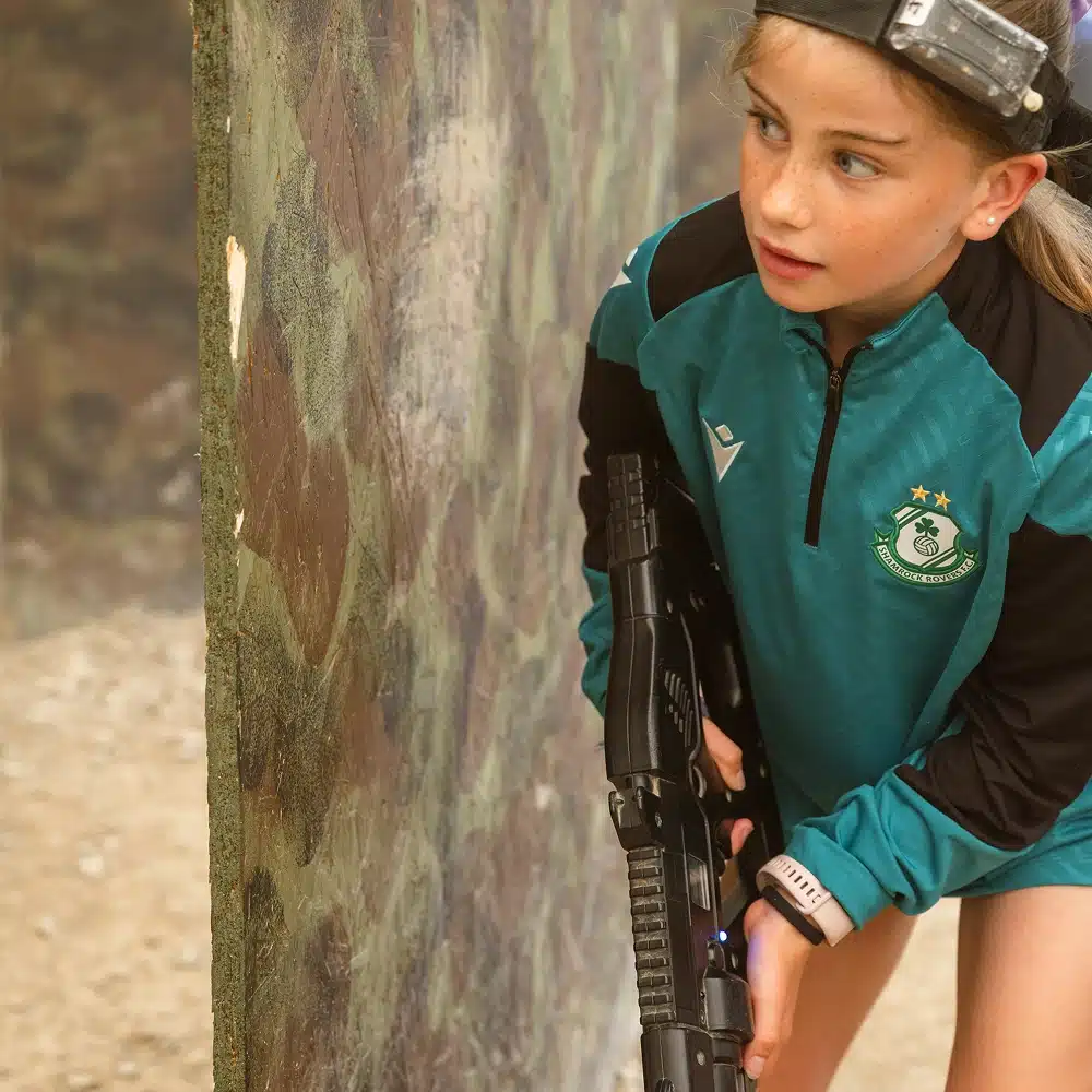 A young girl wearing a green sports jersey and headband holds a laser tag gun while hiding behind a camouflaged wall at a Navan laser tag party, appearing focused and ready to play.