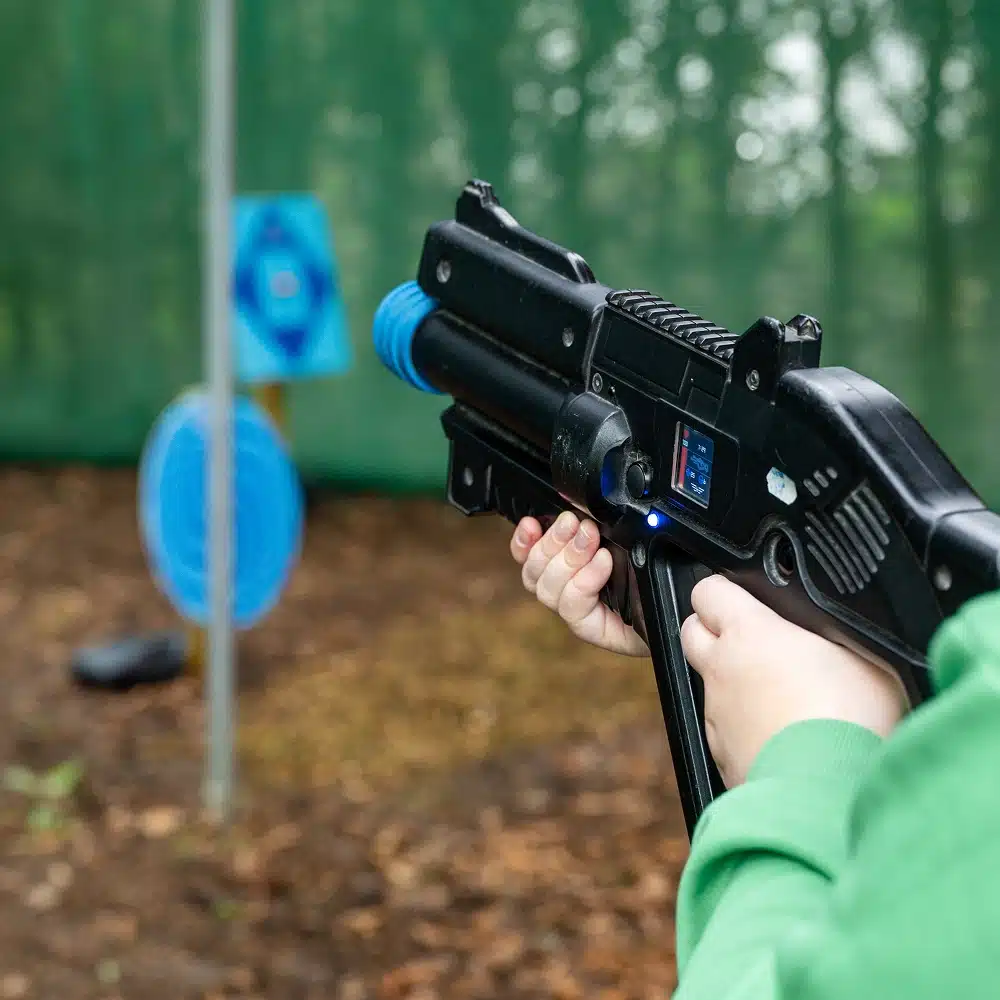 A person in a green sleeve holds a black toy laser gun, aiming at blue circular targets outdoors with trees blurred in the background during a Navan laser tag party.