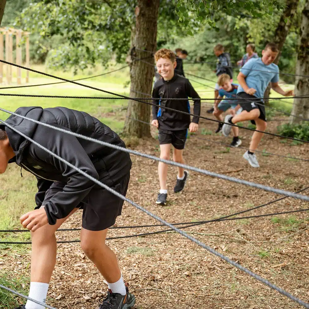 Children navigate an outdoor obstacle course, stepping carefully through ropes tied between trees on a grassy area, much like the thrill of a Navan laser tag party, with more children in the background enjoying the activity.