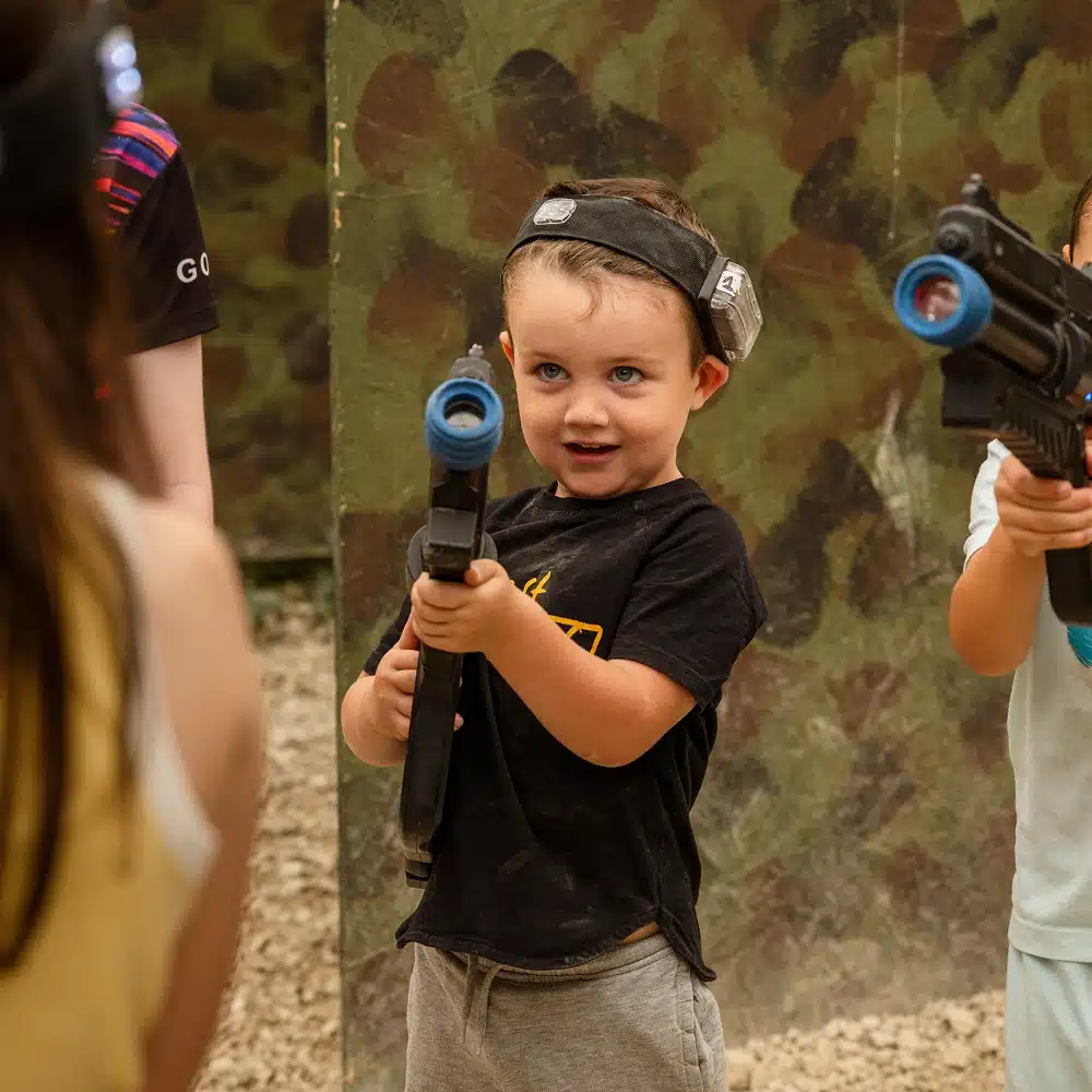 A young child wearing a headband and holding a toy laser gun stands outdoors, smiling and aiming forward during a Navan laser tag party, with another child and adult nearby in a play area with camouflage walls.