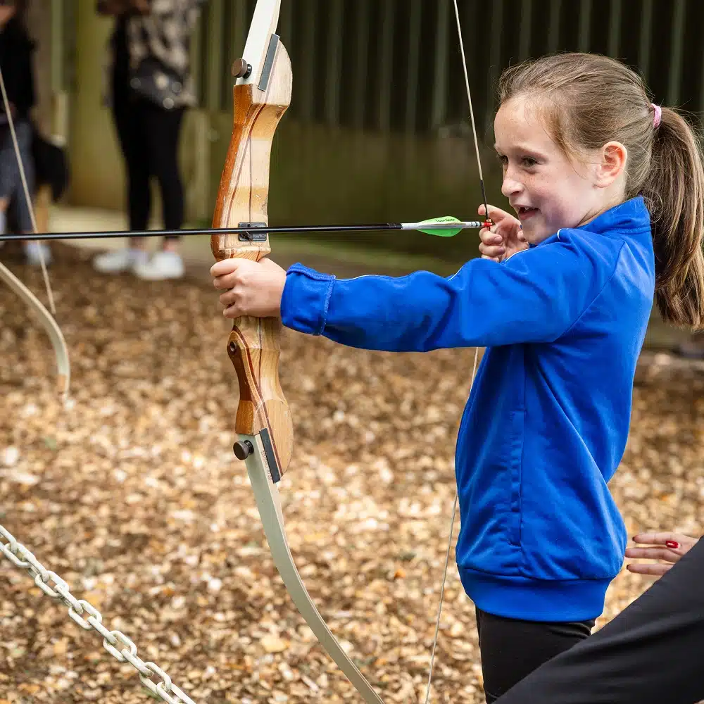 A young girl in a blue jacket smiles as she draws a bow and aims an arrow at an archery range during a Navan laser tag party, with people and greenery blurred in the background.