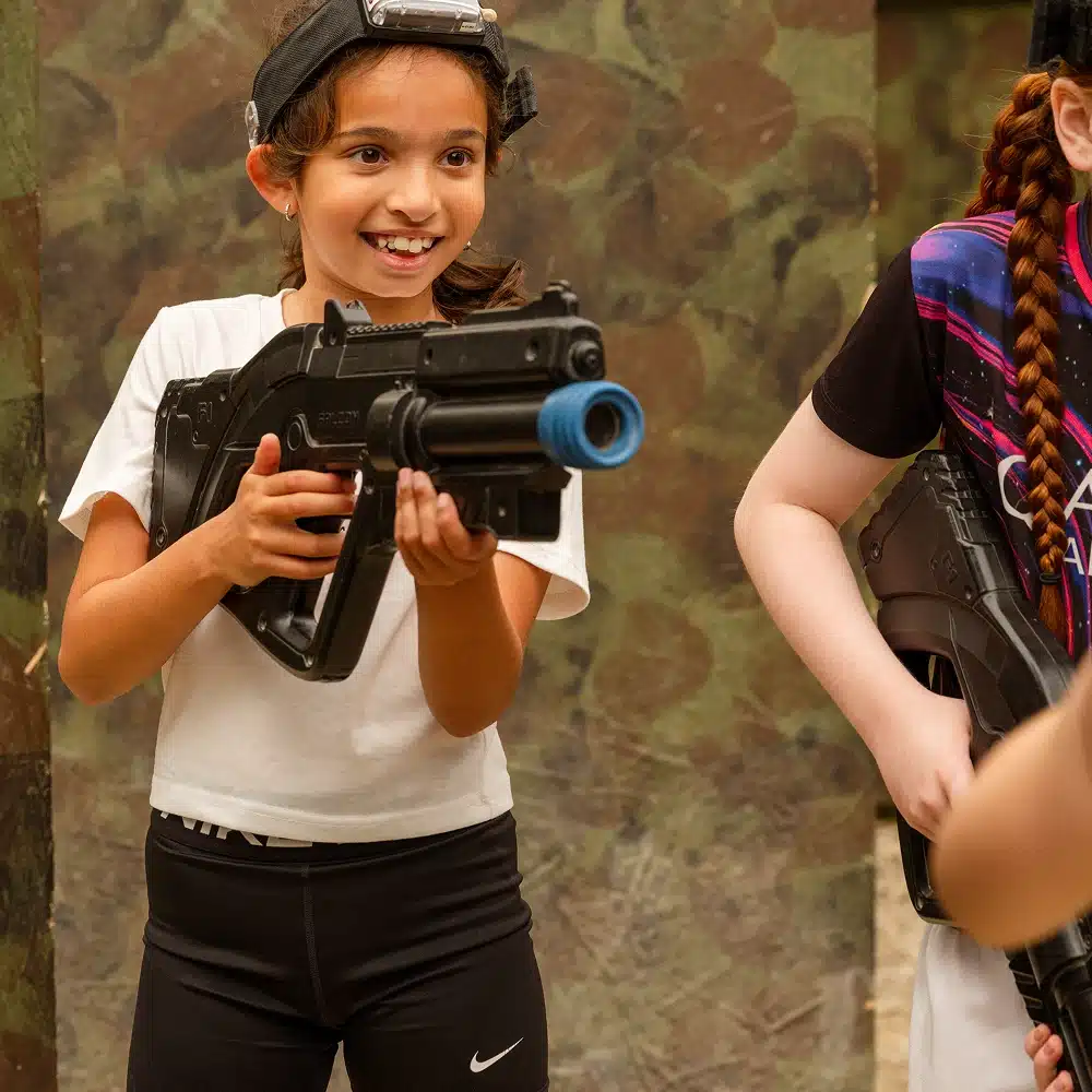 A smiling young girl wearing a headband and white T-shirt holds a large toy gun, playing laser tag at a Navan laser tag party indoors with a camouflage wall in the background. Another child is partially visible beside her.