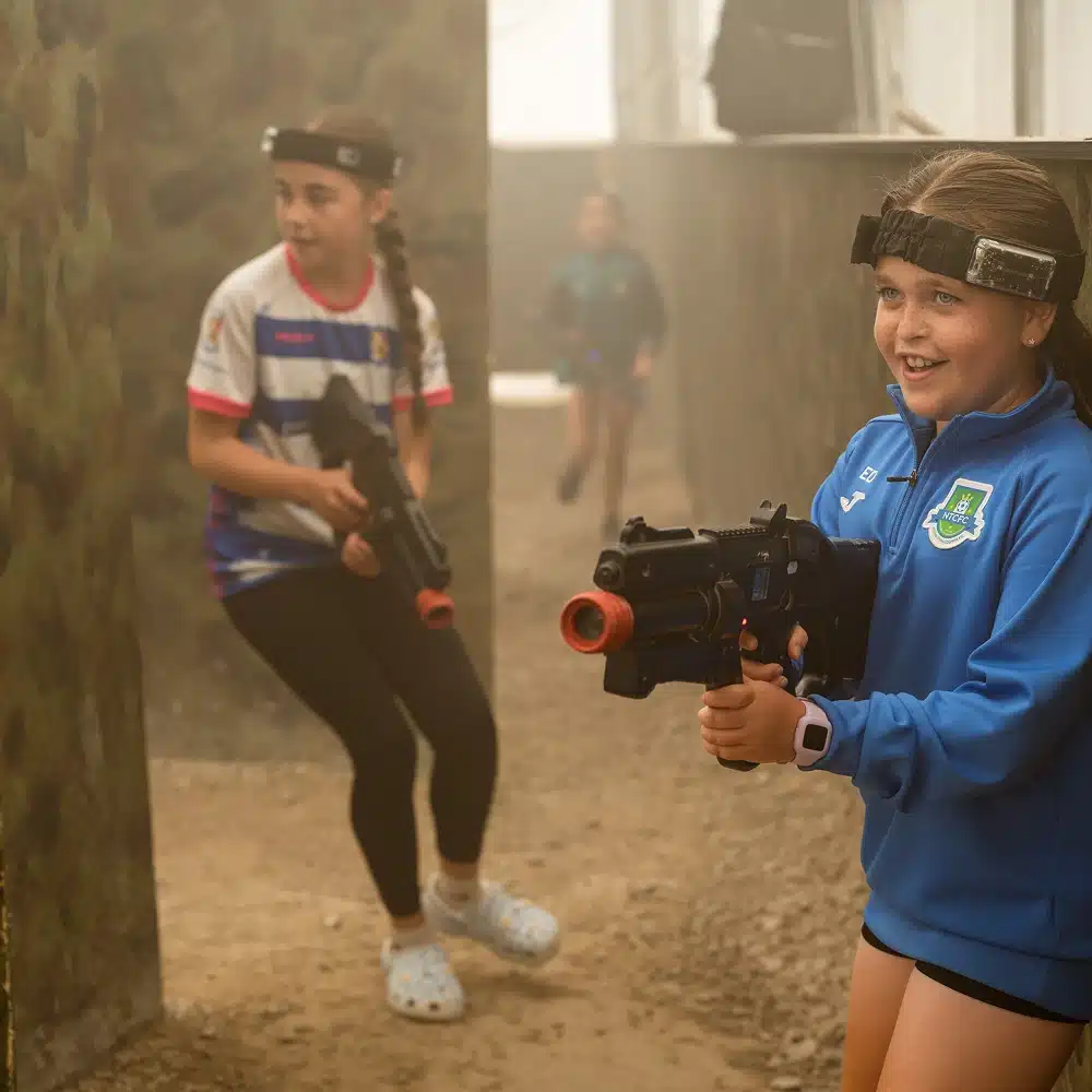 Two girls wearing hairbands and holding toy laser tag guns play in an outdoor arena with camouflaged walls. Both appear focused and excited as they move through the space at a thrilling Navan laser tag party.
