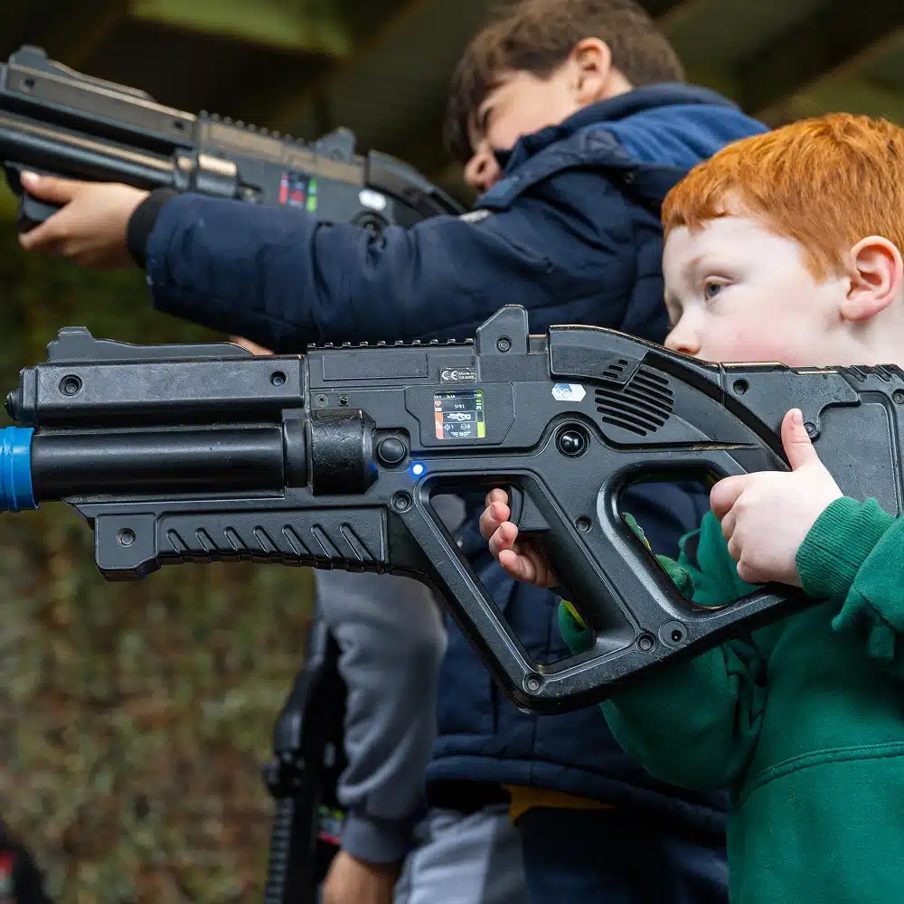A young boy with red hair aims a large toy laser gun at a Navan laser tag party, with another child holding a similar toy in the background. Both are outdoors, dressed warmly, focussed on a game.