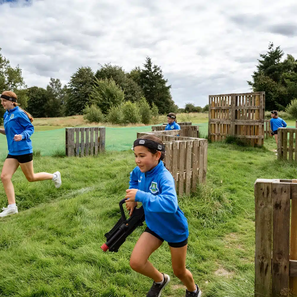 Children in blue sweatshirts play an outdoor Navan laser tag party game, running between wooden barriers on a grassy field under a partly cloudy sky. One child in the foreground holds a laser gun and looks focused.