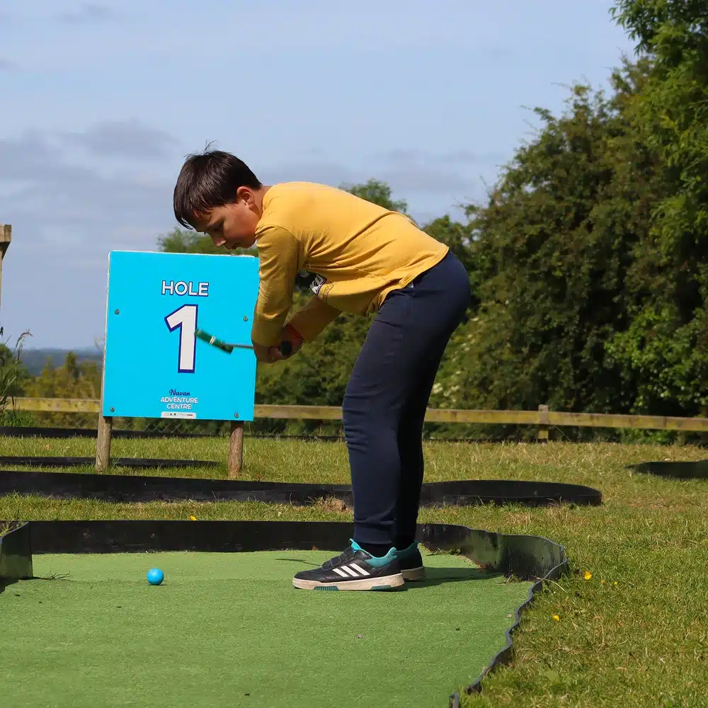 A young boy in a yellow shirt and navy trousers prepares to hit a blue ball with a mini-golf club at hole 1 on an outdoor mini-golf course, perfect for a fun day out or even a Navan laser tag party. Trees and blue sky are in the background.