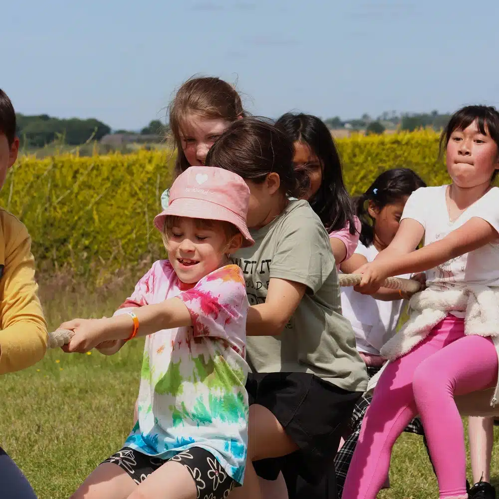 A group of children outdoors enthusiastically playing tug-of-war, pulling on a rope together during a Navan laser tag party. The kids look determined and are enjoying the sunny day, surrounded by green grass and bushes.