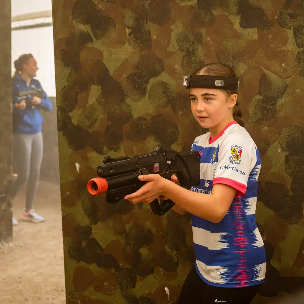 A girl in a striped sports jersey and laser tag headband crouches with a laser tag gun, aiming round a camouflaged barrier at an exciting Navan laser tag party. Another player is visible in the background, partially obscured by mist.