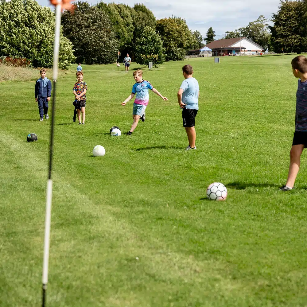 A group of children from Navan primary school play football on a grassy field, with one child kicking a ball whilst others watch nearby. Trees and a building are visible in the background, capturing the fun before their laser tag activity.