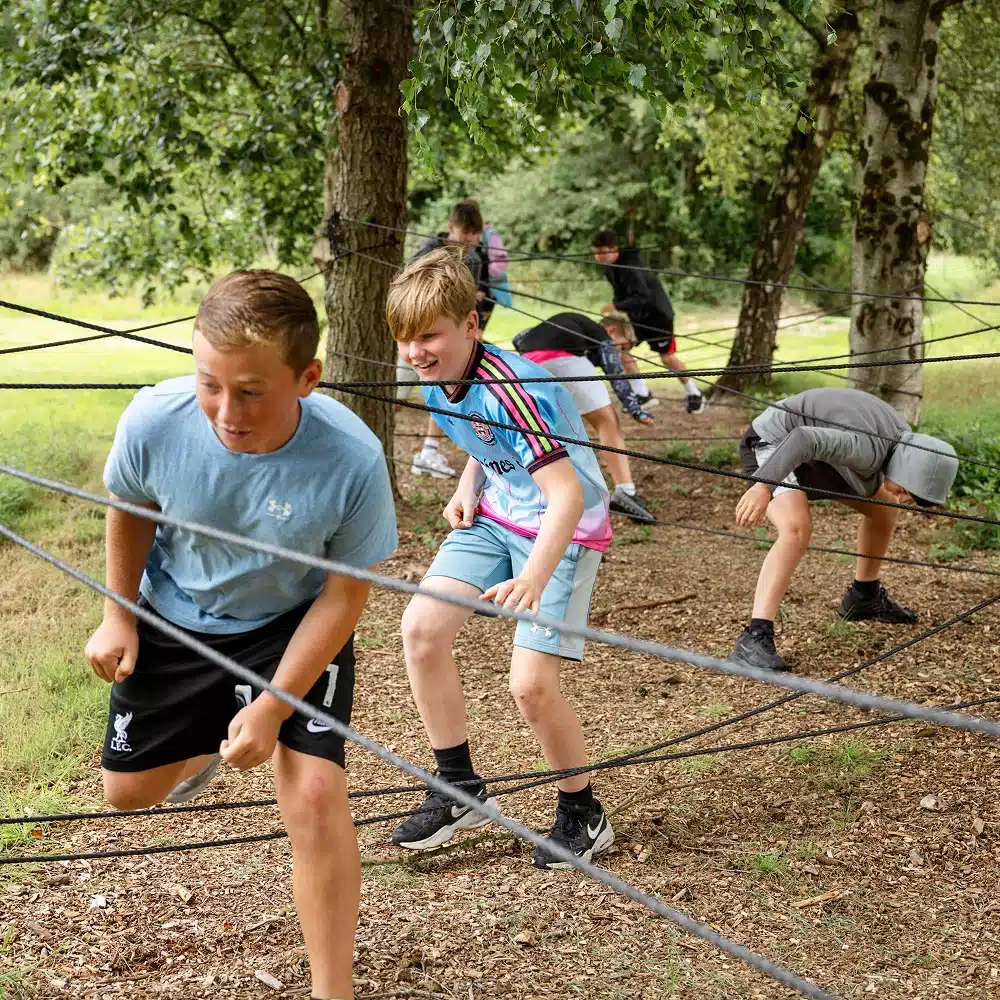 A group of boys from Navan primary school are outdoors, navigating through a web of black ropes tied between trees, bending and ducking as if playing an adventurous game of laser quest in a wooded area.