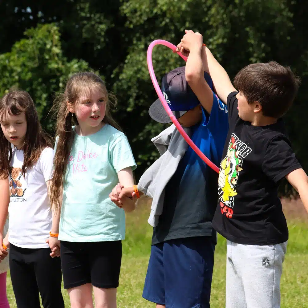 Four children stand outside on grass at Navan primary school, holding hands. One boy passes a pink hula hoop over his head and arm whilst others watch, waiting their turn. Trees and sunlight brighten the scene—perfect for outdoor fun like laser tag.